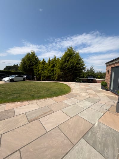 The image shows a paved patio area with various-colored stones, bordered by a semi-circular patch of green grass. Lush green trees and a section of a building are visible in the background. The sky above is blue with scattered clouds. A white car is partially visible near the trees. The overall scene is an outdoor setting on a sunny day.