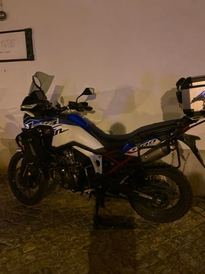 A parked motorcycle, a Honda Africa Twin, is positioned against a white wall on a cobblestone street in Tavira, Portugal. The motorcycle is predominantly white and blue with red accents and features a large windscreen, side panniers, and a top box. The wall behind the motorcycle has a framed sign on the left side with text that reads "mas" and some smaller text below. The ground is composed of textured cobblestones, and the lighting suggests it is either dusk or night, with artificial light illuminating the scene. There are no people visible in the image.