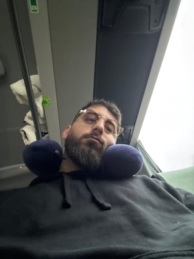 An adult male is centrally depicted, seated within the interior of what appears to be a passenger transport vehicle. He possesses dark hair, a full beard, and wears gold-rimmed eyeglasses along with a dark-colored hooded sweatshirt. A navy blue U-shaped neck pillow is positioned around his neck. His gaze is directed towards the camera with a neutral to slightly narrowed expression. To his left, a light-colored, possibly padded object is visible hanging near a grey interior panel, which features a circular green sticker with indecipherable markings. The vehicle's ceiling is grey, with an overhead fixture present. To the right, an overcast sky is visible through a window, and a segment of a green upholstered seat or headrest is partially depicted. The male is seated; no other individuals or direct interactions are visible. There are no specific visual indicators within the image to confirm the location as Rosny-sur-Seine, France.