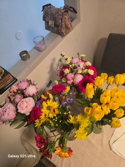 This image depicts a still life of several bouquets of flowers on a light-colored tablecloth, with a glimpse of an upholstered chair and a wall in the background. The flowers include vibrant yellow tulips, pink and white peonies, fuchsia gerbera daisies, yellow chrysanthemums, and some smaller accent flowers in shades of blue and red. A decorative handbag with a leopard-print pattern rests on a white shelf or ledge above the flowers, alongside a lit candle in a glass jar and a metal object that appears to be a candle holder. A trophy or award plaque is also visible on the shelf, engraved with text including "BEAUTY JUDGING ACADEMY" and a name. The scene is lit by natural light, creating soft shadows. The lower right corner of the image displays the text "Galaxy S25 Ultra." There is no information available about the location in the city of Vancouver, United States of America, in this image.