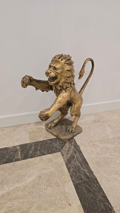 A life-sized, golden-colored statue of a lion is positioned on a marble floor in a white-walled room. The lion is depicted in a dynamic pose, with its mouth open in a roar, claws extended, and body contorted as if in mid-leap or charge. Its tail curls upwards. The floor is made of large, light-colored marble tiles with darker marble accents forming a geometric pattern. The statue is resting on a rough, irregular base. The lighting appears to be artificial and even, casting minimal shadows. The overall scene suggests an indoor setting, possibly a lobby or a display area. The location is identified as Kazafani, Northern Cyprus / Cyprus.