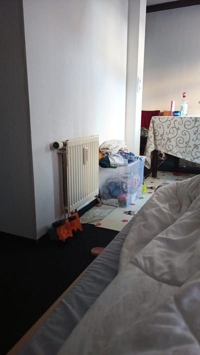 The image depicts an indoor scene, possibly a bedroom or living area, with elements suggesting a lived-in environment. In the foreground, a bed with white comforters dominates the lower right portion of the frame, partially obscuring the floor. The floor is covered with a dark carpet. To the left, a white radiator is mounted on a textured off-white wall. In front of the radiator, on the floor, are several colorful children's toys, including orange and green building blocks. To the right of the radiator, a large clear plastic bin overflows with laundry, including various colored clothes. A pale rug with a pattern of cartoon animals is visible beneath the bin and toys. Further to the right, a table covered with a white cloth featuring an ornate swirling pattern is partially visible. On the table, there are several bottles, including one with a red cap that appears to be a cleaning product or cosmetic. The overall lighting suggests an indoor setting with natural light entering from an unseen source. There are no people visible in the image. The scene is consistent with a domestic environment in Altdorf bei Nürnberg, Germany.