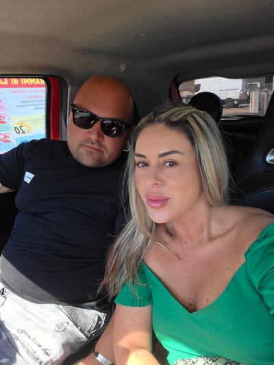 This is a selfie taken inside a vehicle, likely a car, with two people. The image is oriented vertically. A woman with blonde and brown hair is leaning in towards the camera, with her face close to the lens. She is wearing a green top that reveals her décolletage. A subtle silver piercing is visible on her collarbone. Next to her, a man with a shaved head and sunglasses is also looking towards the camera. He is wearing a black t-shirt and what appears to be distressed denim jeans. Behind them, part of the car's interior is visible, including black leather seats and the window. Outside the window, a street scene can be glimpsed, suggesting they are in an urban or semi-urban environment. A sign with Italian text and pricing information is partially visible on the left side of the frame, with legible text including "Jib IMMAЯ" and "€ 50". The lighting suggests it is daytime. The overall mood of the image appears casual and intimate.