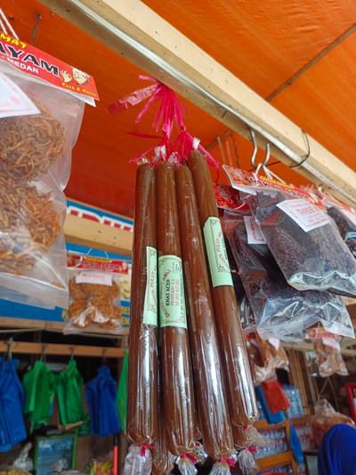 The image shows a close-up view of various food items, likely for sale, hanging from a wooden structure above an orange awning. The items include several long, brown, cylindrical snacks wrapped in clear plastic, possibly a type of sweet confection. There are also bags of dried food products, some appearing to be crispy snacks, and some dark, leafy-looking items. Several of the packaged items have labels with text, including "DODOL PULUT ASLI KHAS ACEH" and "AYAM". In the background, other merchandise and some brightly colored fabric items are visible. The setting appears to be a market stall. The presence of "ACEH" on a label strongly suggests a connection to the province of Aceh, Indonesia. Given the context of food stalls and local products, Sawang, a regency in Aceh, is a plausible location within Indonesia where such a scene might be found.