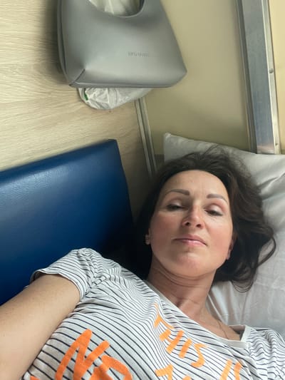 A woman lies on a bed in what appears to be a train compartment. She is wearing a striped t-shirt with orange lettering. To her left is a blue padded surface, likely a seat or bunk. Above her is a light grey handbag with the word "BLUMI" visible on it, along with a white plastic bag. The walls of the compartment are light wood-paneled and a pale yellow color. A white pillow is visible behind her head. The image does not provide any information about the specific location within the city of Babino 2, Russia.