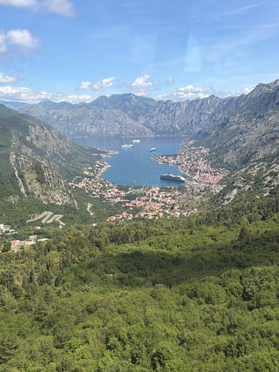 An aerial, high-angle panoramic view captures the stunning natural and urban landscape of Mirac, Montenegro, likely overlooking the Bay of Kotor. The scene is illuminated by bright daylight under a clear blue sky dotted with scattered white clouds.

In the center of the image, a deep blue body of water, resembling a fjord or bay, curves through rugged, grey-green mountains that rise majestically from its shores and form the background. Along the waterfront, a densely built town stretches, characterized by numerous buildings with distinctive red and orange tiled roofs, indicative of Mediterranean architecture. Several boats are visible in the bay, including a large white cruise ship seemingly anchored or docked near the town, and smaller vessels.

To the right, a vast, dense green forest covers the steep mountain slopes, extending into the foreground and showcasing varied shades of green foliage. Winding switchback roads, carved into the forested mountainside, are visible on the right, suggesting pathways leading up or down the terrain. The overall setting portrays a harmonious blend of natural grandeur and human settlement, with the serene bay and the dramatic mountains under a sunny sky. No visible text is present.