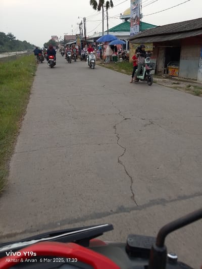 The image shows a street in Kecamatan Gabuswetan, Indonesia. The street is paved with asphalt, which is cracked. Several people are riding motorcycles, traveling in the same direction. There are buildings on both sides of the street and a few people are standing in the foreground. The image is taken from a motorcycle, with the handle bar and some of the red body of the motorcycle visible in the foreground. The sky is cloudy and the image was taken on March 6, 2023 at 5:20 pm.