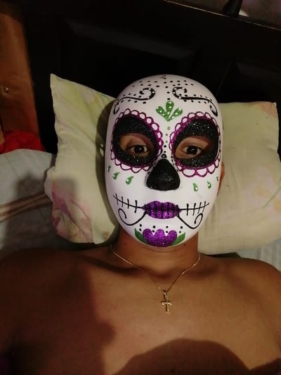 A person lies in bed wearing a sugar skull mask. The mask is white with black eye sockets outlined in purple glitter, a black nose, and purple glitter lips with stitched detail. Green glitter accents the forehead. A gold cross necklace is visible on the person's bare chest. A pillow is behind their head. The scene is dimly lit.
