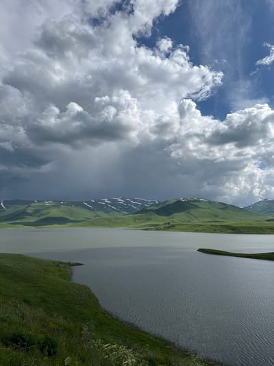 The image depicts a serene landscape featuring a tranquil lake in the foreground, with rolling green hills and snow-capped mountains in the background. The sky is a dramatic canvas of contrasting clouds, with patches of blue sky visible amidst heavy, dark storm clouds on the left and lighter, fluffy white clouds on the right. The lake's surface is rippled, reflecting the sky. The foreground shows a grassy embankment leading down to the water. The location is identified as Tsghuk, Armenia, suggesting a natural, mountainous terrain. No people or specific actions are visible, creating a sense of stillness and natural beauty.