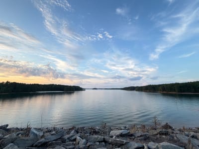 A wide-angle, eye-level, outdoor photograph captures a serene view of Lake Lanier in Buford, Georgia, at sunset. The foreground is dominated by a rocky shore, with various-sized gray and tan stones scattered across the frame. Dry grass and small twigs are visible amongst the rocks. The middle ground features the expansive, calm surface of Lake Lanier, reflecting the soft hues of the sky. Gentle ripples disturb the water's surface, creating horizontal lines that extend across the lake.
The background is defined by a dense, dark green treeline that lines the opposite shore, interrupted by a few distant islands. The horizon is clearly visible, separating the lake from the sky. The sky above is a canvas of blue, interspersed with wispy white and gray clouds. Towards the left side of the frame, near the horizon, the sky shows hints of orange and yellow, indicating the setting sun. The overall impression is one of tranquility and natural beauty at dusk. There are no people or animals visible in the image.