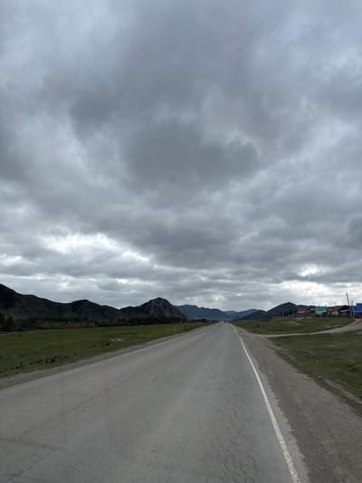 The image shows a gray asphalt road with a white dividing line, stretching into the distance. The road is flanked by green grassy fields and mountains. The sky is overcast with a heavy layer of gray clouds. In the distance, some buildings can be seen along the road. The location is Ust-Kan, Russia.