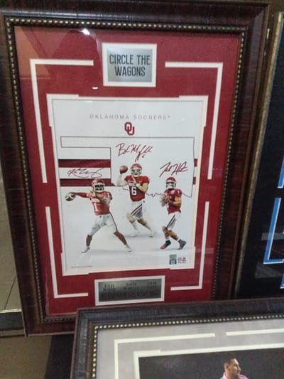 A framed Oklahoma Sooners memorabilia piece features an image of three football players, Kyler Murray, Baker Mayfield, and Jalen Hurts. Each player's signature is printed above them. "Circle the Wagons" is written in a box at the top. The frame has a brown wooden finish with red and white matting. A nameplate at the bottom identifies the players and the "Franchise Foundation." A portion of another framed item sits below with a similar frame.