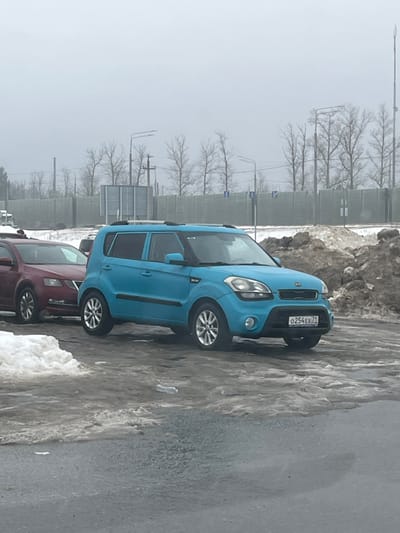 This image depicts an outdoor scene in Sholokhovo, Russia, under an overcast sky. The primary subject is a light blue (cyan) first-generation Kia Soul, bearing Russian license plate `O254EX71`. The vehicle is parked on a wet, dark asphalt or tarmac surface, alongside a partial accumulation of dirty snow and slush. To the left and slightly behind the Kia, a dark red sedan, possibly a Skoda Octavia, is partially visible. To the right of the Kia, a large mound of dirty snow mixed with earth and debris is present. The immediate foreground shows patches of melting snow and standing water on the dark pavement, with a clear plastic bottle lying horizontally. In the background, a light grey sound barrier wall extends horizontally, behind which a line of bare deciduous trees, utility poles, and streetlights are visible against a uniformly pale sky. No individuals are present in the frame.
