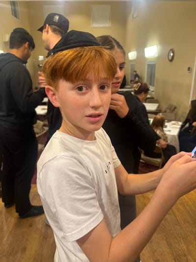 A young boy with red hair and a black yarmulke looks at the camera. He wears a white t-shirt. A woman with braided hair stands behind him. In the background are people sitting at tables. The room has a ceiling with lights and a wooden floor. Someone holds a phone up, suggesting they are taking a picture or video.