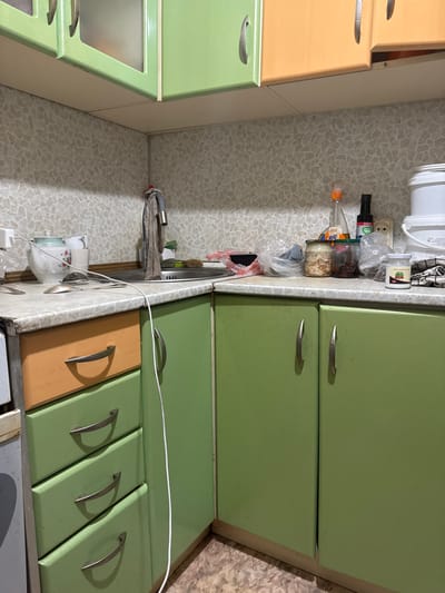 This image depicts a cluttered kitchen counter and cabinets in a residential setting. The cabinets are painted in two shades: a pale orange on the upper section and a light green on the lower section, with silver handles. The countertop is a speckled grey laminate.
On the counter, to the left, a white teapot with a floral design and a white cup are visible. A sink with a faucet and a dishtowel hanging from it is located towards the back. Various items are scattered around the sink area, including plastic bags containing food items, two glass jars with contents, a bottle of soy sauce, a bottle of cooking oil, and a small jar with a label that appears to be in Cyrillic. To the far right, a large white bucket or container is present. A white electrical cord runs diagonally across the scene.
The backsplash is a beige wallpaper with a mosaic-like pattern of irregular grey and white tiles. The floor is tiled with a light-colored material featuring a speckled, irregular pattern.
The image does not provide any information that can definitively place it in the city of Демихово, Russia, or any other specific location. The scene appears to be a typical, lived-in kitchen environment. There are no people visible in the image, and no specific actions or interactions are depicted. The lighting is somewhat dim, suggesting it might be indoors with artificial lighting.