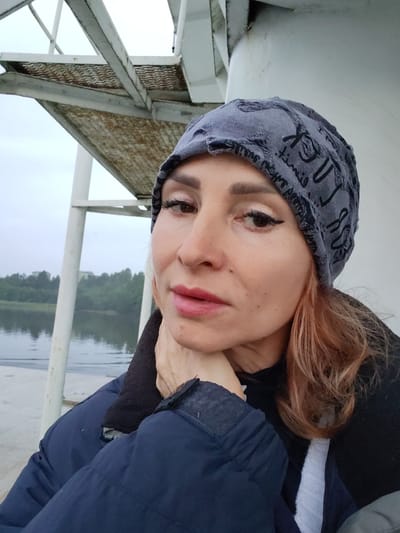 A medium shot captures a woman wearing a grey beanie and a dark blue jacket, positioned in the foreground against a backdrop of a cloudy sky and a body of water with a treeline. The woman is looking towards the camera with her right hand resting on her chin. Her hair, a shade of reddish-brown, is visible on the right side of the frame. In the background, a white structure with metal stairs is visible to the left, and a body of water is seen with trees on the opposite bank. The location is identified as Ivanov Bor, Russia.