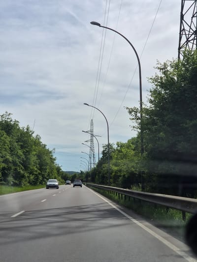 The image depicts a highway scene near Sanem, Luxembourg. The view is from within a moving vehicle, looking down the road. Several cars are visible ahead. On either side of the road, there are dense trees and bushes, creating a green barrier. Tall, arched street lights are positioned along the roadside, and behind them, there are electrical towers. The sky above is partially cloudy. A guardrail runs along the right side of the road. The road itself is gray with white lane markers.