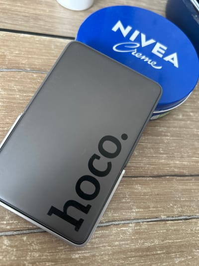 A dark grey rectangular object with the brand name "hoco." printed in black lettering is placed on a light brown wooden surface. To the right of the object, a blue circular tin with "NIVEA Creme" printed in white lettering is partially visible. The wooden surface has a visible grain pattern and appears to be a table or floor. There are no people in the image. The scene is indoors, likely in a domestic setting. The city of Kokhma, Russia is not mentioned or depicted in the image.