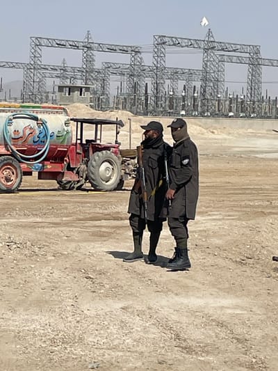 The image depicts an outdoor scene in Pir Dust, Afghanistan, featuring two armed individuals standing on barren, light-brown, rocky ground. In the background, a large electrical substation with numerous gray metal lattice structures, insulators, and transformers extends across the horizon. A small, light-colored building with a dark roof is visible within the substation complex, and a small white flag or banner is affixed to one of the taller structures.
To the left of the two individuals, a red tractor is parked, pulling a light blue water tanker trailer adorned with colorful Farsi/Dari script and decorative elements.
The two individuals are positioned in the foreground, facing slightly right. Both wear dark, uniform-like clothing and black baseball caps. The individual on the left has a dark scarf covering the lower part of their face and carries a rifle, likely an AK-47 variant, slung across their body. They wear dark pants and dark-colored footwear. The individual on the right wears a light tan or olive-colored balaclava covering their lower face and carries an object, possibly another rifle or piece of equipment, slung across their body. They wear dark pants and dark combat boots. Shadows are cast by the individuals, indicating direct sunlight from an elevated source. The sky is clear and pale blue.