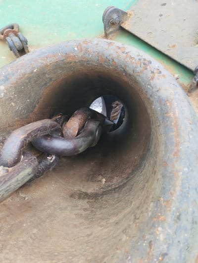 The image captures a close-up, downward perspective of a heavy-duty maritime feature, likely on a ship or in a port setting in Городище, Russia. The central subject is a large, conical metal hawsepipe or chocks, through which a robust anchor chain passes.

The hawsepipe itself is made of dark, weathered metal, showing significant signs of age and corrosion. Its interior and exterior surfaces are covered in pitted rust and a faded, chipped grey-blue paint. The anchor chain, composed of thick, dark grey links, is heavily rusted, particularly in its recesses and where the paint has worn away. Several links are visible, coiling into the hawsepipe and leading into the unseen depths.

Surrounding the hawsepipe is a flat, metal surface painted a muted, faded green. This surface also exhibits considerable wear, with patches of rust and discolored areas, indicating prolonged exposure to weather and salt. In the upper right of the frame, parts of the ship's structure are visible, including bolted fasteners that secure a metal plate.

Through the opening at the end of the hawsepipe, a distant and obscured background is visible, suggesting an open environment, possibly water or a dock. The scene is brightly lit, indicating daytime. The textures are predominantly rough and corroded, highlighting the industrial nature of the equipment and the harsh conditions it endures. The image portrays a static moment; no direct activity is taking place. There is no visible text.