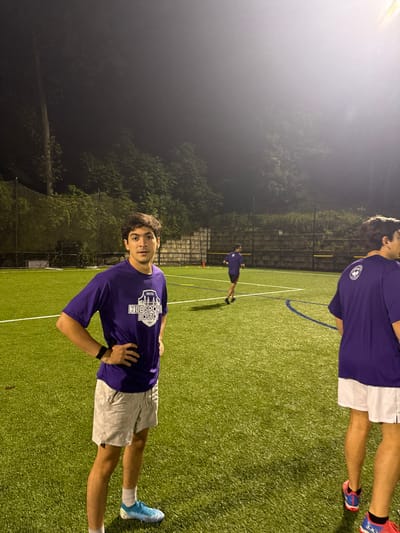 The image depicts an artificial turf sports field at night, illuminated by overhead floodlights. Three individuals are present on the field, which features white and blue line markings.
In the foreground, slightly to the left, a young male stands facing the camera. He has dark, wavy hair and a neutral expression. He wears a short-sleeved purple t-shirt with "HUBSPORTS BOSTON" and a building-like logo printed in white on the chest. He is also wearing light grey athletic shorts, white socks, and light blue athletic shoes, possibly turf cleats. His hands are placed on his hips, and a dark-colored fitness tracker is visible on his left wrist.
In the midground, positioned towards the center-right, another young male in a purple t-shirt and dark shorts is observed jogging away from the camera. His back is mostly visible as he moves across the field, with small orange cones present on the turf in his vicinity.
Partially visible in the foreground, on the right side of the frame, is the lower torso and legs of a third young male. He wears a purple t-shirt with a white circular logo on the back, white shorts, and brightly colored athletic shoes (pink/red and blue). He appears to be standing still, facing away from the camera.
The background consists of dark, dense foliage, suggesting a wooded area or park border. A dark fence structure runs along the perimeter between the field and the foliage, with a concrete or block retaining wall visible behind it in some sections. The scene's overall lighting indicates an evening or nighttime sports activity.
This scene is located on an artificial turf field in Newton, Massachusetts, United States.
