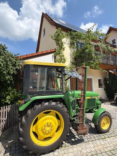 A John Deere 1040 tractor is parked on a cobblestone driveway in front of a yellow house with solar panels on the roof in Ebermannstadt, Germany. The tractor is green and yellow, with large rear tires and a smaller front tire. A green hedge is to the left of the tractor, and a wooden fence is behind it. The house has a balcony with plants and a vine growing on the side. The sky is blue with white clouds.