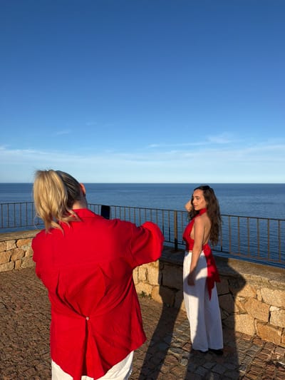 In Santa Maria Navarrese, Italy, two women are captured in a photograph on a cobblestone terrace overlooking the sea. A woman with blonde hair pulled into a ponytail, wearing a red shirt and white pants, is taking a picture with her smartphone. Facing away from the camera, she is positioned in the foreground. To her right and slightly further back, a woman with long, dark curly hair is posing for the photo. She is wearing a red halter-neck top with a tie detail in the back and flowing white pants. The background features a clear blue sky meeting the calm, blue sea, separated by a metal railing. The stone wall of the terrace is visible in the lower part of the image. The lighting suggests it is late afternoon, casting distinct shadows on the ground.
