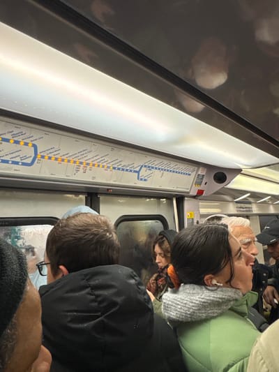 A slightly tilted view inside a crowded train. People are standing close to each other, their faces and bodies partially visible. A map showing the train route is affixed to the wall. The map includes the names of stations such as Mitry-Claye, Vert-Gatant, Sevran-Livry, Parc des Expositions, Villepinte, and Bramy. The reflection in the window indicates a rainy day.