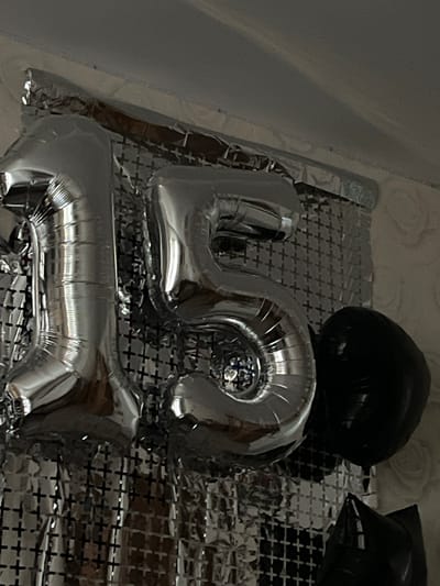 The image shows a close-up of silver foil balloons shaped as the numbers "1" and "5," indicating the number fifteen. They are positioned against a backdrop of silver, crinkled material with a grid pattern. To the right of the "5" balloon, a large, round black balloon is visible. Partially visible at the bottom right is a black, pointed object, possibly another balloon or a decorative element. The lighting suggests an indoor setting, likely a celebratory decoration, and there is no visible connection to Novoanninskiy, Russia.