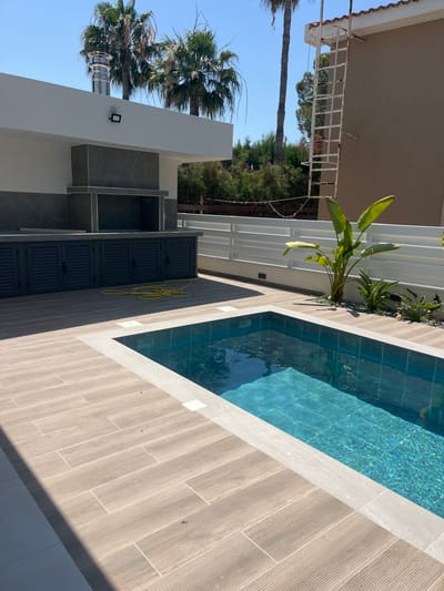 The image depicts an outdoor residential area in Moni Community, Cyprus, featuring a newly constructed swimming pool and an adjacent outdoor kitchen. The pool is rectangular with blue tiled water and a built-in step. The pool is surrounded by light-colored tiled decking. To the left of the pool is an outdoor kitchen area with a modern design, including dark grey cabinetry with louvered doors, a dark stone countertop, and a built-in grill area with a stainless steel chimney extending upwards. A yellow garden hose is coiled on the deck in front of the cabinetry. Behind the kitchen and pool, a white horizontal fence separates the property from other areas. In the background, there are palm trees, dense green foliage, and a light brown building with a white metal ladder attached to its side. The sky is clear and blue, indicating a sunny day. There are no people visible in the image.