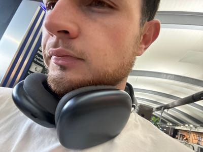 A close-up, low-angle shot captures the lower half of a man's face and head, with large black headphones positioned around his neck. The man has short, light brown stubble on his chin and jawline, and his lips are slightly parted. He is wearing a white t-shirt. In the background, a curved ceiling with white and gray stripes suggests an indoor public space, possibly an airport terminal. To the left, a tall, narrow object with blue and white vertical stripes is visible. The overall setting appears to be a modern, well-lit environment. The location is identified as Apodaca, Mexico.