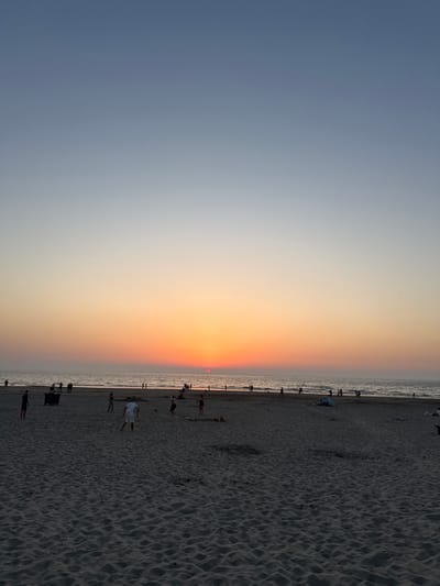 The image depicts a serene beach scene at sunset in the city of Hook of Holland, Netherlands. The sky transitions from a pale blue at the top to vibrant hues of orange and pink near the horizon, where the sun is setting over the sea. The water is calm, with gentle waves lapping the shore. The sandy beach is populated by numerous individuals engaged in various activities. In the foreground, several people are visible on the sand. Further back, closer to the waterline, groups of people are strolling, playing, or simply relaxing. One group appears to be playing soccer, with a ball visible in the sand. A few beach umbrellas and a trash bin are also present on the beach. The overall atmosphere is peaceful and recreational, characteristic of a popular beach destination during the evening.