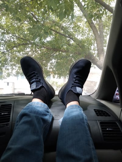 The image shows a first-person perspective of a person's legs and feet resting on the dashboard of a vehicle, with the legs covered by blue jeans. The person is wearing dark blue athletic shoes with black socks. Through the windshield, a tree with green leaves is visible, along with a building and parts of the sky. The interior of the car, including the dashboard and air vents, is also in view. The scene appears to be outdoors during the daytime.
