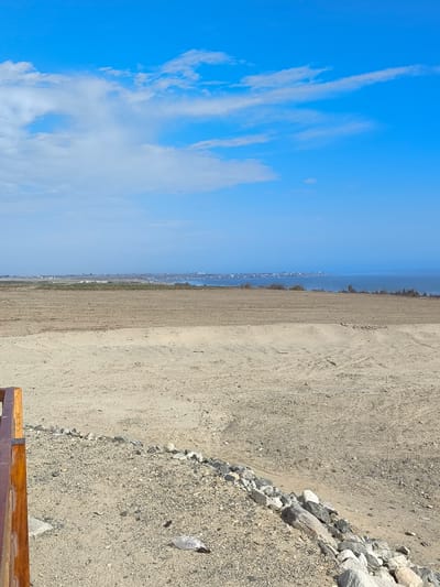 The image captures a coastal view in Huanchaco, Peru. A clear blue sky with scattered white clouds dominates the upper portion of the frame. Below, a light-brown, sandy landscape extends toward the horizon. In the mid-ground, there is a darker, plowed field. A thin, hazy line indicates the shoreline with faint buildings of the city visible along the coast. A wooden structure is partially visible in the lower left corner. In the foreground, the ground is littered with small rocks and a single plastic bag, indicating some level of human presence and potential littering. The light suggests it is daytime, with clear visibility.