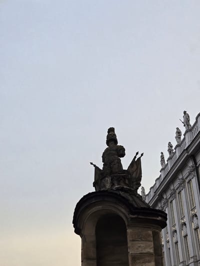 This image is an upward-angle shot of the exterior of a building with architectural details, specifically gargoyles and ornate carvings. In the foreground, a large, dark, stone gargoyle is prominently featured. It appears weathered and menacing, with sharp angles and a gaping mouth. Behind and above it, a series of smaller, lighter-colored figures, also possibly gargoyles or statues, line the facade of the building. These figures are more detailed, with what appear to be human-like forms dressed in robes, some holding objects that could be swords or staffs. The building itself is grand, with a patterned ceiling or frieze visible above the figures. The sky is overcast and pale, suggesting it could be daytime with cloudy weather. The overall impression is one of ancient architecture, possibly a cathedral or a historic public building. The orientation of the image is unconventional, with the building appearing to hang from the top of the frame. No text is visible in the image. The location context is Ansbach, Germany, which aligns with the Gothic or Neo-Gothic architectural style suggested by the gargoyles and ornate stonework.