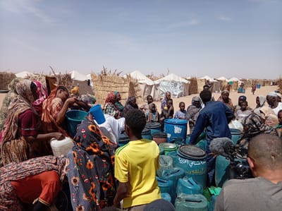 The image captures an outdoor scene in what appears to be a displacement camp or temporary settlement in Goshabi, Sudan, under a bright, clear blue sky. The ground is dusty and arid, with rudimentary shelters made of dried straw or reeds interspersed with white canvas tents in the background.

A large group of people, including men, women, and children, are gathered in the foreground and midground. Most of the women are wearing headscarves and traditional clothing. The central activity involves the collection or distribution of water. Numerous blue and green plastic barrels and smaller jerry cans are densely packed together, and individuals are seen handling these containers, some actively pouring water using blue plastic jugs and buckets.

The scene is brightly lit by the sun, suggesting midday or early afternoon. In the immediate foreground, with his back to the viewer, a person is operating a professional video camera fitted with a shotgun microphone, indicating the presence of media or a documentary crew. Arabic script is visible on some of the blue barrels; specifically, one large blue barrel on the right prominently displays "منظمة تشادي الخيرية" (Chad Charity Organization), with "عائشة و..." (Aisha and...) written below it. The overall impression is one of a community managing essential resources in challenging conditions.
