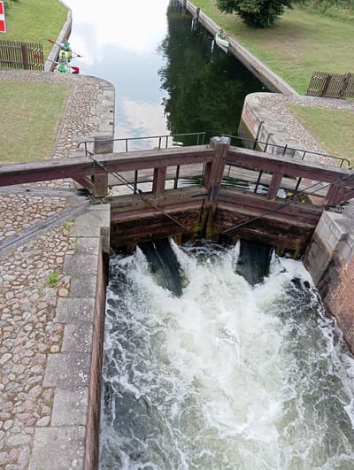 In Mikaszówka, Poland, a canal lock is in operation, with water cascading forcefully from two sluices into a lower section of the waterway. The lock gate is made of weathered brown wood, with metal reinforcements and cables visible. The surrounding banks are lined with stone and cobblestones, and a grassy area with a wooden fence is present to the left. Two people in kayaks, wearing life vests and holding paddles, are positioned in the upper section of the canal, to the left of the lock mechanism. One kayak is directly behind the other, and the individuals appear to be waiting. The water is a deep green, reflecting the sky and surrounding trees.