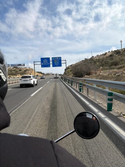 The image presents a first-person view from a motorcycle traveling on a multi-lane asphalt highway. The rider's black helmet is partially visible in the upper left foreground, and a circular rearview mirror reflecting dark clothing occupies the lower right foreground. The highway, identified as the A-31, features two lanes separated by a broken white line and a solid white line marking the right shoulder. Dark linear tire marks are present on the road surface.
To the right, a metal guardrail with green and white striped cylindrical bollards and orange reflectors lines the road, bordering a dry, scrub-covered hillside. An electricity pylon with power lines stands on the upper part of this hillside. In the distance, on the left side, another hillside with sparse vegetation and a billboard is visible.
In the midground, an overhead gantry displays two blue directional signs. The left sign indicates "A-31 Alacant Alicante aeropuerto" with a straight-ahead arrow. The right sign, marked "1550 m," directs traffic to "N-330 Aspe" and "E-15 A-7 Elx / Elche Murcia" with a diagonal right arrow. This specific signage places the location on the A-31 motorway in the municipality of Monforte del Cid, Spain, prior to the junction with the A-7/E-15 and N-330, approximately at geographical coordinates 38.3585, -0.6865.
Traffic ahead includes a white Mazda CX-5 SUV with the license plate "2050 NCB" in the left lane, followed by a silver sedan. A white recreational vehicle (RV) is visible in the right lane further ahead. The sky is partially cloudy with scattered white clouds against a pale blue background.