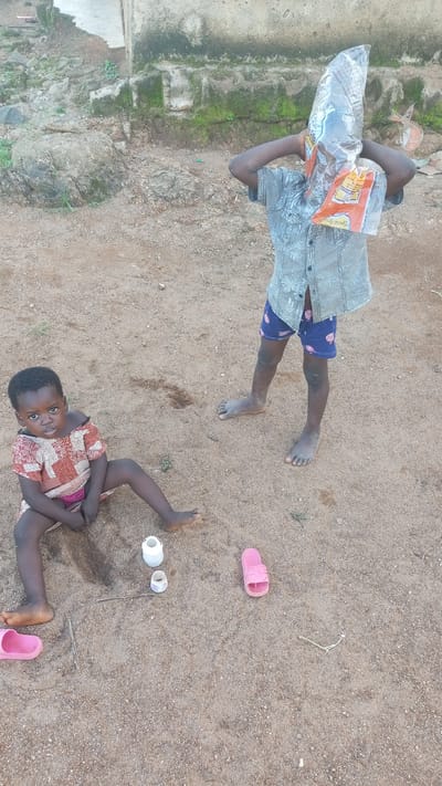 The image depicts two young children in an outdoor, sandy setting, likely in Kagarko South, Nigeria. In the foreground, a young girl with dark skin sits on the sand, facing the camera with a neutral expression. She is wearing a patterned, off-the-shoulder dress and a pink garment underneath. To her left, a pink sandal lies on the ground. In the mid-ground, a second child, possibly a boy, stands with their back to the camera, holding a clear plastic bag over their head, obscuring their face. This child is wearing a patterned short-sleeved shirt and blue shorts with a pattern. A pink sandal is also visible near this child. Scattered on the sand between the children are two small white containers and a thin stick. The background shows a rough, uneven surface with rocks and some vegetation, leading to a weathered, light-colored structure. The lighting suggests it is daytime.