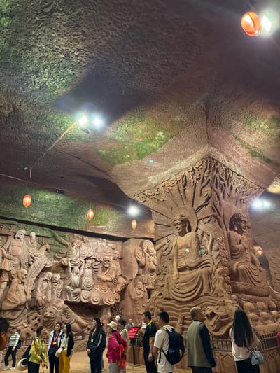 This image captures the interior of the Dazu Rock Carvings, specifically a rock-cut cave, located in the city of Shizhong, China. The cave walls are covered in detailed carvings depicting Buddhist figures. The ceiling is rough and uneven, with patches of moss and is lit by both white and orange lights. A group of people are standing near one of the carved walls, appearing to observe and admire the artwork. The lighting conditions are dim overall.