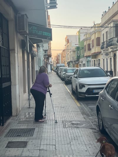 The media file depicts a street scene in La Línea De La Concepción, Spain. The main subject is a woman using a crutch for support as she walks on a tiled sidewalk. She is wearing a purple shirt and black pants. In the background, several cars are parked along the street, including a white Kia. Buildings with multiple stories line the street, displaying a variety of architectural styles and colors. A sign for "the STYLIST STATION by EVA" is visible on one of the buildings, along with a phone number: 956 17 54 25. The time of day appears to be late afternoon or early evening, with soft, warm lighting suggesting the golden hour. The weather seems clear. A brown dog on a leash can be seen in the upper right portion of the image. The scene conveys a sense of everyday life in a Spanish town.