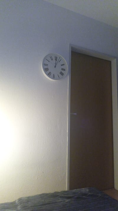 The image presents an indoor scene, likely a room in a residential building located in Ginsheim-Gustavsburg, Germany. A prominent feature is a round, white wall clock with black Roman numerals mounted on a light-colored wall. The clock's hands indicate the time is approximately 12:10 or 12:11.

To the right of the clock, a dark brown door, possibly wooden, is partially visible, featuring a metallic handle. The left side of the room, particularly the wall below and to the left of the clock, is brightly illuminated by a strong, localized artificial light source, creating a stark contrast with the dimmer lighting in the rest of the frame. In the bottom foreground, a dark, textured surface, consistent with a bed covered by a blanket or duvet, is present.

The scene is static, devoid of people or activity, conveying a sense of quiet and stillness. The combination of artificial lighting and the time displayed on the clock suggests the photograph was taken either late at night or very early in the morning. The visible text consists solely of the Roman numerals (I through XII) on the clock face.
