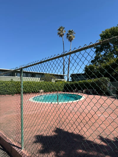 The image shows a fenced-in area with a pink wall and a turquoise oval pool. The fence is made of chain-link and has a green metal frame. The wall has a textured surface and is partially covered in shadows. There are palm trees visible in the background, as well as green foliage growing along the wall. The sky is a clear blue, suggesting it is daytime and the weather is sunny. The scene appears to be outdoors, possibly in a residential or recreational setting. No people are visible in the image, and no text is present. The overall impression is one of a private, serene outdoor space. The location is identified as San Mateo, United States of America.