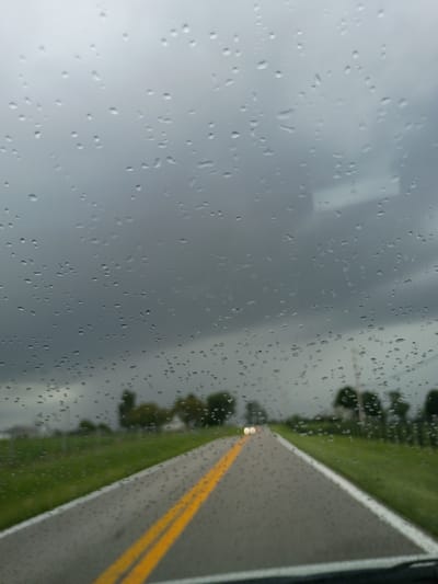 This image captures a view through a car windshield on a rainy day. Water droplets are visible on the glass, distorting the scene outside. The road ahead is paved and has two yellow lines down the center. To the sides of the road are grassy areas with scattered trees and some houses in the distance. The sky is overcast with dark, stormy clouds. The overall impression is one of driving through inclement weather in a rural or suburban area. The location is not specified as Shelbyville, United States.