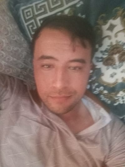 A male individual, appearing to be a young adult with short dark hair and light facial stubble, is depicted in a close-up selfie. He is wearing a light-colored, possibly beige or light grey, long-sleeved garment with a subtle, textured pattern of fine horizontal or diagonal lines. A white collar or undershirt is visible at the neck opening on the right side. The individual has dark eyes and a neutral expression with a slight smile, gazing towards the camera. The background consists of two distinct patterned fabrics: a lighter grey fabric with a swirling, curvilinear design on the left, and a darker teal or blue-green fabric featuring a mix of floral and geometric fret patterns on the right. The scene indicates an indoor setting in the city of Pichakchi, Uzbekistan.