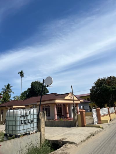 This is an outdoor, eye-level, full-shot photograph taken in Dalam, Indonesia. The scene depicts a street with a building and a large water tank on the right side, and a clear blue sky with wispy white clouds on the left. The building features a yellow facade with pink accents and a brown roof, and has a decorative fence in front of it. The water tank is a large white cube with a metal cage, and appears to have black graffiti on its side. The time of day is likely daytime, given the bright lighting and clear sky. The weather appears to be sunny and clear. There is no visible activity or event taking place, and no people are present. The image evokes a sense of a quiet, residential street in a tropical location. The graffiti on the water tank is partially visible and reads "LELA".
