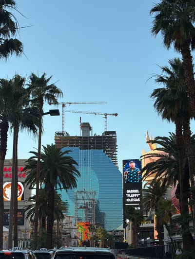 The image shows a street scene in Paradise, Nevada, USA, with palm trees framing the view. A building under construction with two cranes is visible in the center of the frame. A large LED screen displays an advertisement for Gabriel "Fluffy" Iglesias. A line of taxis is parked on the right side of the street.