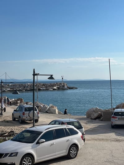 The image depicts a scene at the harbor in Potos, Greece, under a clear blue sky. The foreground features a white station wagon parked on a paved surface, with another silver station wagon visible behind it, partially obscured. A silver SUV is parked further back, facing away from the camera, with a spare tire mounted on its rear. Several large rocks form a breakwater along the edge of the harbor. In the middle ground, the blue water of the sea is visible, meeting the rocky breakwater. Further out, a distant hazy landmass is visible on the horizon. Two streetlights with dark, angled shades are positioned along the walkway, and a thin, tall pole stands vertically on the right side of the image. A person is seated on the rocks overlooking the sea. A small lighthouse-like structure is visible on the breakwater in the distance. Several people are walking on the walkway to the left, near a trailer.