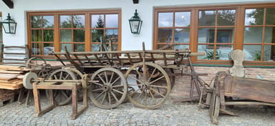 The image depicts an outdoor scene in front of a building with white walls and large, multi-paned wooden-framed windows. The windows reflect trees and the sky, indicating an overcast day. Two green, traditional lantern-style lights are mounted on the wall.
In the foreground, a collection of antique agricultural equipment is displayed on a cobblestone ground. Prominently featured is a wooden wagon with large spoked wheels and a ladder-like structure. To the left of the wagon, there is a wooden frame holding a grinding stone, with visible gears and a crank mechanism. Stacked lumber is leaning against a wooden rack to the far left. To the right of the wagon, another piece of wooden agricultural machinery, possibly a threshing machine, is situated. A unique wooden object with a circular top and a narrower base, resembling a stylized human figure, stands upright between the wagon and the machinery on the right.
The overall impression is that of an outdoor exhibit of historical farm implements, possibly at a museum or a traditional establishment in Eugendorf, Austria. No people are visible in the image. The scene is static and focused on the arrangement of these antique objects.