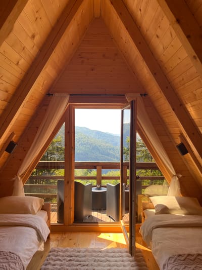 The image depicts the interior of a wooden A-frame cabin with a panoramic view of lush green mountains. Sunlight streams into the room, illuminating the wooden paneling of the walls and ceiling. Two beds with white bedding are positioned on either side of the room, facing a large glass door that opens onto a balcony. The balcony features two dark chairs and a small round table. The view through the glass door reveals a dense forest covering rolling hills under a bright sky. The overall impression is one of tranquility and natural beauty. The location is specified as Katun Kobil do, Montenegro.